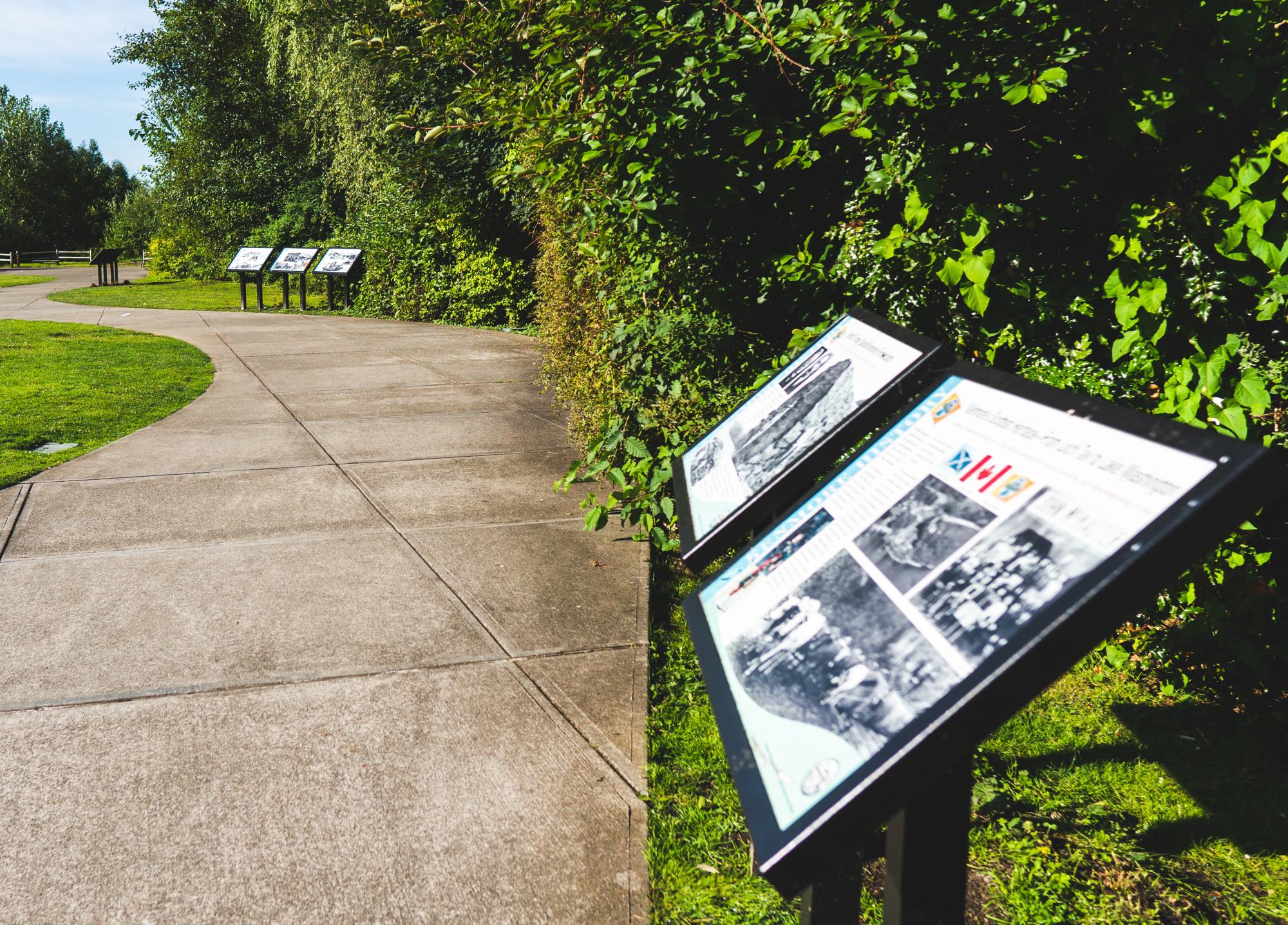 Historical signs at Log Boom Park along a nice, paved walking path on a bright sunny day. The text of the signs are not visible. 