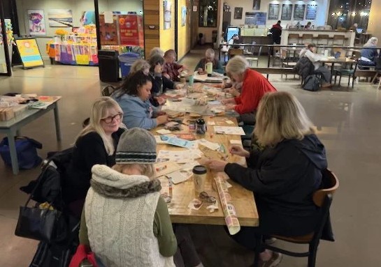 A group of individuals gather around a table inside the Hangar to work collaboratively on an art piece.